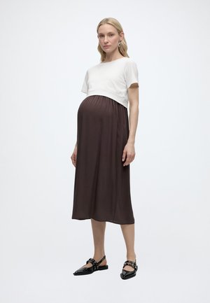Femme enceinte debout, portant un haut blanc à manches courtes sur une robe midi marron, avec des ballerines noires pointues sur un fond uni.