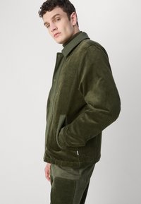 Olive green corduroy jacket with a collared neck, front pockets, and textured fabric. Smooth finish, subtle branding on the chest.