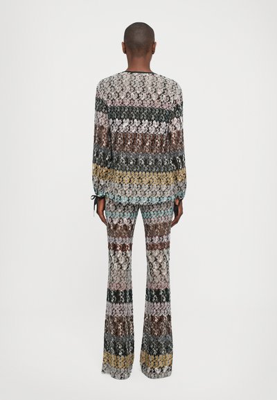Person with short hair stands facing away wearing a multicolored knit sweater and matching flared pants against a white background.