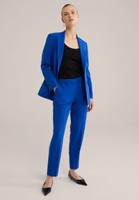 Ryškiai mėlynas pritaikytas kostiumas su struktūrine švarku ir siaurais kelnių modeliais, derinamas su juodu trumpu topu ir juodais smailiais kulnais.