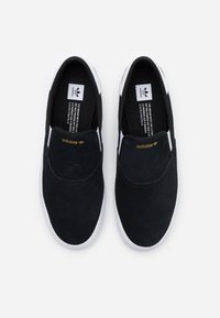 Ett par svarta Adidas slip-on sneakers med vita sulor och en guldlogotyp på ovansidan, sedd ovanifrån på en vit bakgrund.