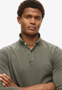 Camisa verde de manga larga con una tapeta de botones y cuello de solapa. La tela parece suave con una textura lisa. Los brazos del modelo están cruzados.