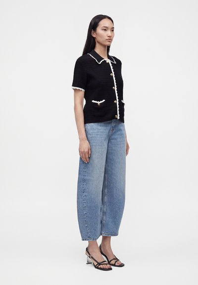 Femme debout portant un cardigan noir à manches courtes boutonné avec bordure blanche, un jean bleu à jambes larges et des sandales à talons noires à brides.