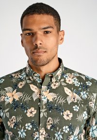Camicia a maniche corte verde oliva con bottoni, decorata con vari fiori e foglie multicolori, colletto strutturato e tessuto morbido.