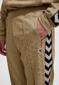 Beežid dressipüksid peene mesilase mustriga, külgmise tõmblukuga tasku, musta ja beeži värvi ketrajaga triip ning väike mesilase logo tasku lähedal. Käsi taskus.