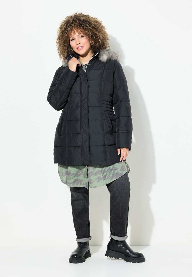Chaqueta de plumas negra con capucha de piel sintética, diseño acolchado, bolsillos laterales y corte recto. Llevada sobre un conjunto en capas en verde y gris.