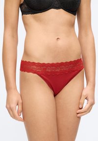 Ženska v črnem čipkasti modrček in rdečih spodnjicah s čipkastim robom in pasom z blagovno znamko, stoji pred enakomerno svetlo ozadje.