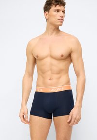 Άνδρας μοντέλο σε φόρμα, φορώντας μπλε ναυτικό μπόξερ Lacoste με πορτοκαλί μέση, στέκεται απέναντι σε ένα απλό ανοιχτό φόντο.