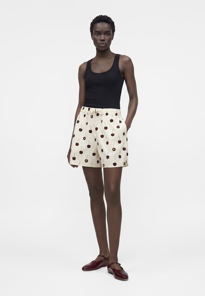 Γυναίκα με μαύρο αμάνικο τοπ, κρεμ σορτς με σχέδιο κερασιών και σκούρα κόκκινα παπούτσια φλατ, στέκεται με το ένα χέρι στην τσέπη.