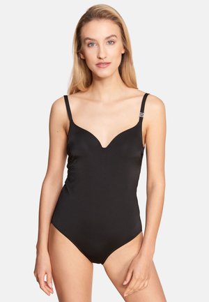 Woman with long blonde hair wears a black fitted one-piece swimsuit with thin straps against a plain light gray background.