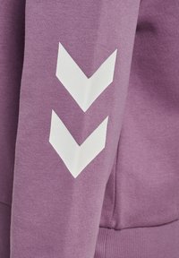 Fioletowy materiał bluzy ze miękką teksturą, z białymi grafikami w kształcie szewronów na rękawie. Prosty, casualowy design odpowiedni na co dzień.