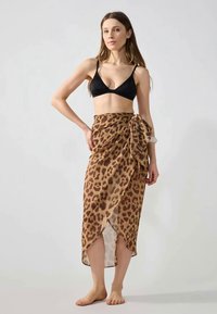 Femme portant un haut de bikini noir et une jupe portefeuille imprimée léopard, debout pieds nus devant un fond clair.