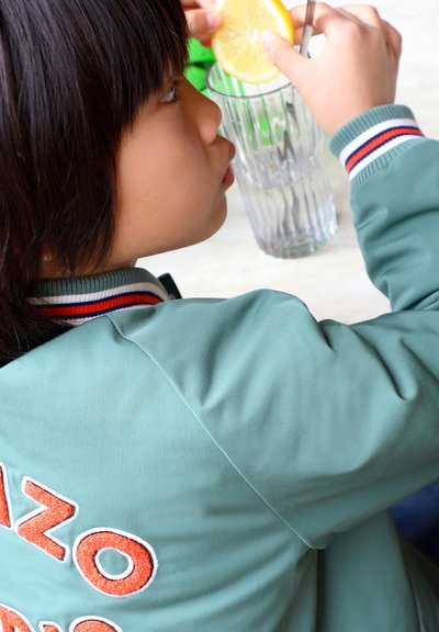 Veste verte en tissu texturé, avec des lettres brodées orange au dos. Un verre avec une tranche de citron est au premier plan.