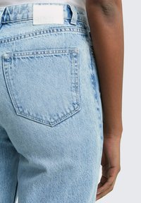 Närbild av en person som bär ljusblå denimjeans med en bakficka synlig, som står med ena armen avslappnad längs sidan.