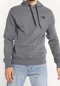 Mężczyzna w szarej bluzie z kapturem The North Face z czarnymi sznurkami i jasnoniebieskich dżinsach, stojący na tle białym.