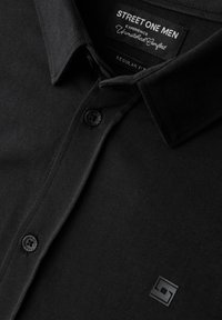 Svart skjorta med knappar och klassisk krage, tillverkad av mjukt tyg, med en liten logopatch och en enda knapp vid kragen.