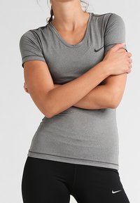 Mujer con los brazos cruzados, vistiendo una camiseta deportiva de manga corta gris de Nike y leggings negros de Nike, frente a un fondo liso.