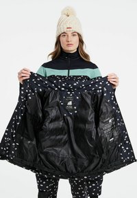 Schwarze Jacke mit weißen Blumendrucken, die das gefütterte Innenfutter und die Etiketten zeigt. Das Modell trägt ein schwarzes Reißverschlussoberteil und eine beigefarbene Strickmütze mit Bommel.
