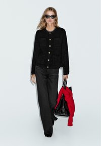 Donna che indossa occhiali da sole neri, cardigan nero con bottoni dorati, pantaloni neri a gamba larga, mentre trasporta una borsa nera con un maglione rosso appoggiato sopra.