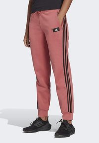 Roze trainingsbroek met zwarte zijkanten, elastische tailleband en een voorzak met een Adidas logo patch. Zacht materiaal en taps toelopend ontwerp.