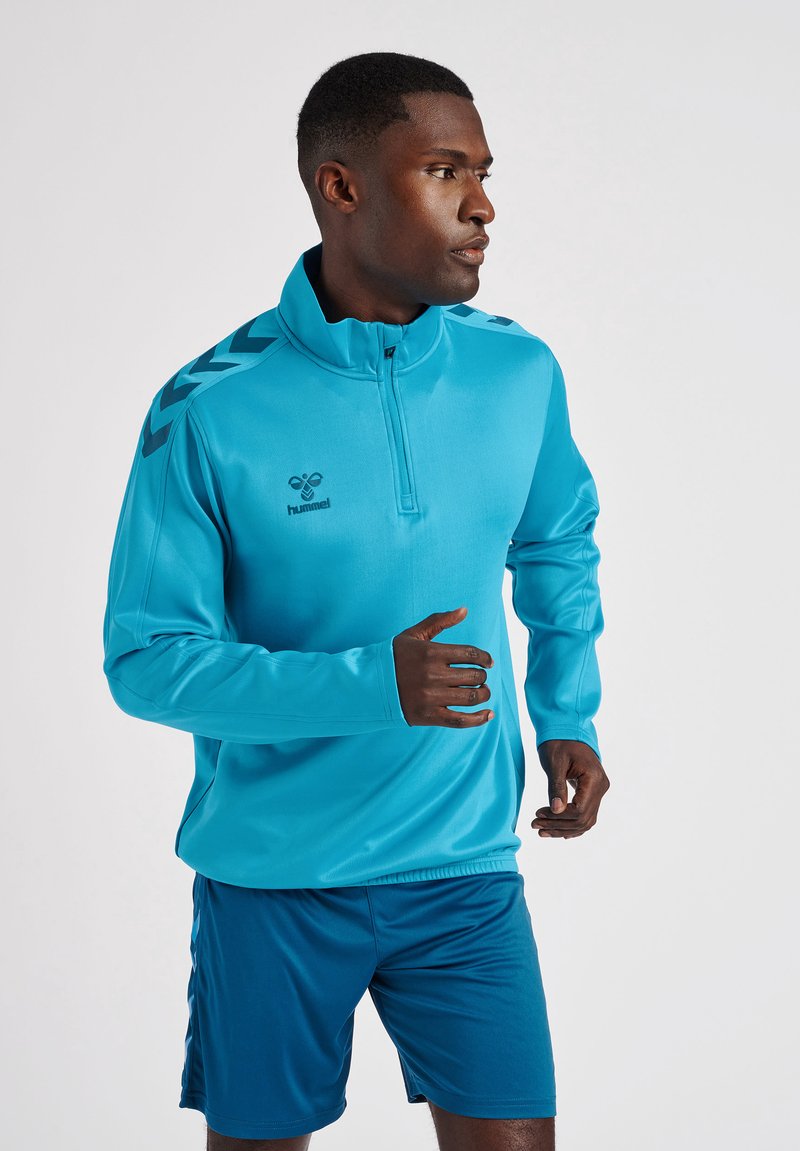 Pullover atletico blu con colletto alto, caratterizzato da accenti neri a zigzag sulle spalle e un logo sul fronte. Indossato con pantaloni corti coordinati.