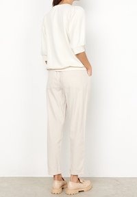 Mujer vestida con pantalones beige claros con bolsillos de parche, una blusa blanca de manga tres cuartos y unos mocasines chunky beige, de pie contra una pared blanca.