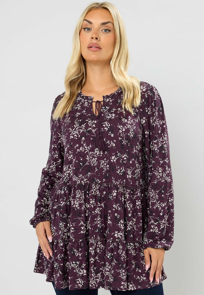 Syklinen tunika syvässä purppurassa, pitkillä hihoilla, rypytetyllä helman reunalla, nyöri-kauluksella, jossa on tupsut, ja valkoisilla kasviaiheetkuvioilla.