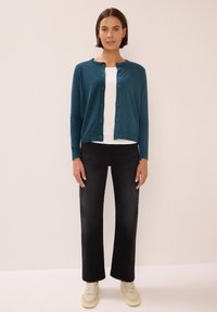 Teal cardigan met ronde hals en knoopsluiting, gelaagd over een witte shirt, gecombineerd met zwarte wijde broek en beige sneakers.