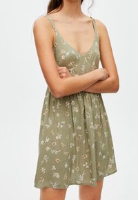 Robe fleurie verte avec un décolleté en V et des bretelles fines. Elle présente de petites fleurs blanches et orange, un tissu doux et un design de jupe évasée.