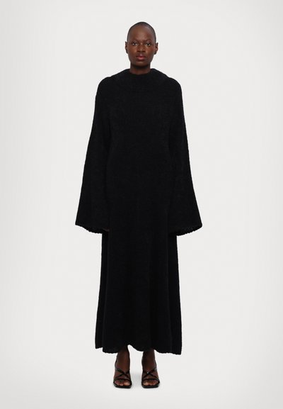 Femme debout portant une longue robe noire en peluche avec de larges manches et des sandales à talons hauts à lanières noires sur un fond blanc uni.