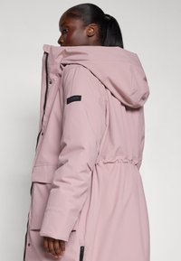 Frau trägt eine locker sitzende, hellrosa Kapuzenparka mit Fronttaschen und schwarzen Reißverschlussdetails, blickt nach links vor einem neutralen Hintergrund.