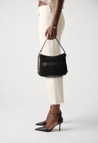 Borsa a tracolla in suede nero con un accento dorato, tracolla a catena e manico texturizzato, tenuta da una persona che indossa pantaloni bianchi risvoltati e tacchi neri.