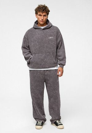 Lichtgrijze, oversized hoodie met een voorzak, gestructureerd materiaal en een wit logo. Gepaard met losse, grijze sweatpants en zwarte sneakers.