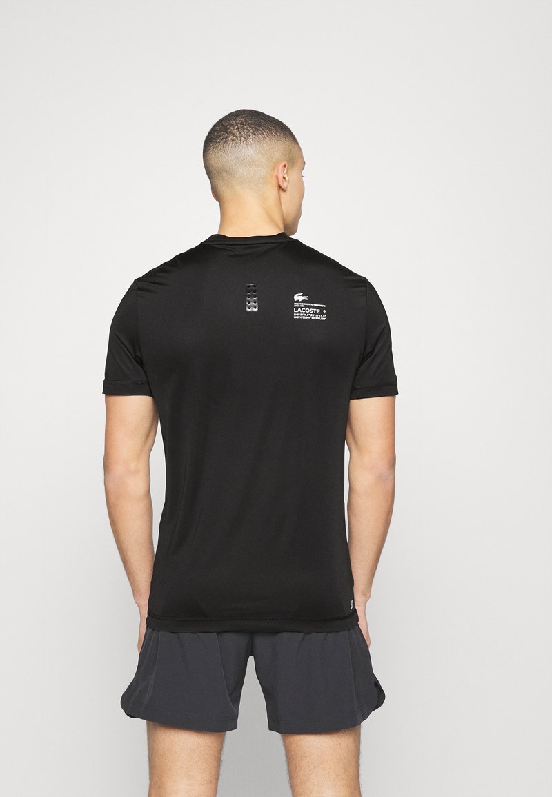 Schwarzes Sport-T-Shirt mit kurzen Ärmeln, mit strukturierten Streifen auf dem Rücken und einem weißen Lacoste-Logo auf der oberen rechten Seite.