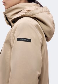 Gros plan d'une personne portant une veste à capuche beige avec un patch rectangulaire noir sur le haut du bras affichant le nom de la marque Icepeak.