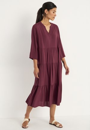 Bordeaux midi-jurk met een V-hals, driekwartmouwen en gelaagde ruches. Soepele stof en een ontspannen silhouet. Draagt met lichte sandalen.