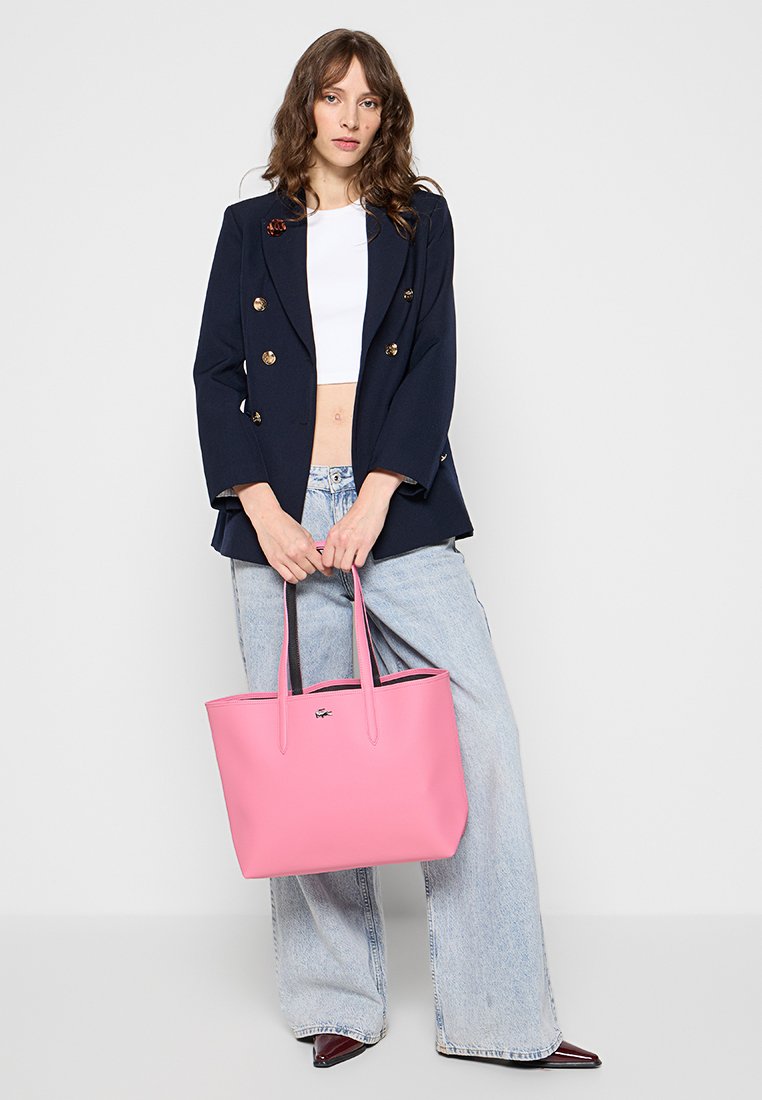Rosa Tote-Bag mit glatter Oberfläche, zwei länglichen Griffen und einem Logodetail, kombiniert mit einem dunklen Blazer und hellblauen weit geschnittenen Jeans.