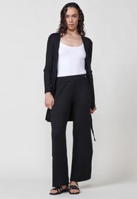 Cardigan negro acanalado de manga larga, usado sobre una camiseta sin mangas blanca. Combinado con pantalones de pierna ancha negros y sandalias de tiras negras.