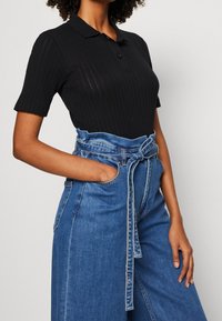 Kvinna som bär en svart ribbad kortärmad polo skjorta nerstoppad i höga blå denimjeans med ett knutet bälte, handen i fickan.