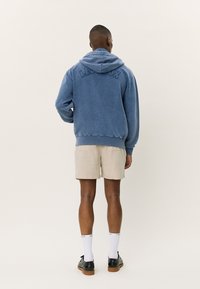Blauwe hoodie met textuurlettering op de rug, relaxed fit en geribde manchetten, gecombineerd met lichtbeige shorts en zwarte schoenen.
