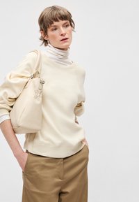 Sudadera color crema con cuello redondo, puños y dobladillo acanalados, combinada con pantalones anchos de pierna en beige. Un bolso beige claro cuelga del hombro.