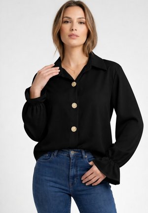 Frau trägt schwarze Bluse mit goldenen Knöpfen und blaue Jeans, posiert mit einer Hand am Kragen und der anderen in der Tasche.