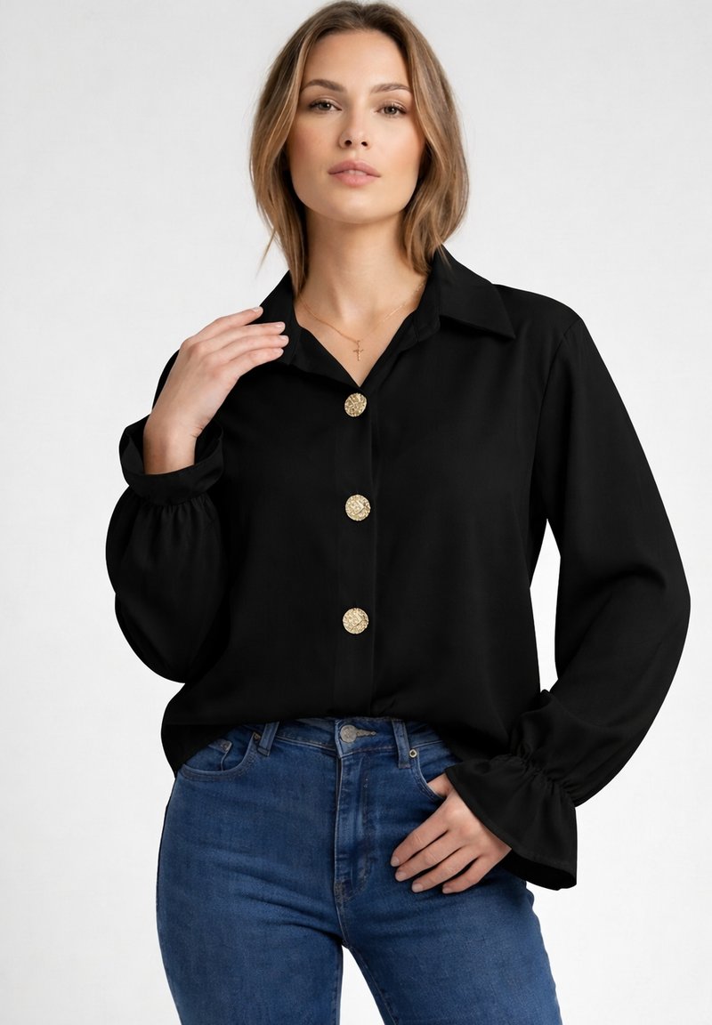Mujer vistiendo una blusa negra con botones dorados y vaqueros azules, posando con una mano cerca del cuello y la otra en el bolsillo.