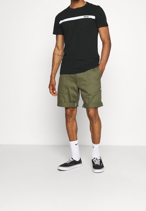 Schwarzes kurzärmeliges T-Shirt mit weißen horizontalen Streifen und Tasche, kombiniert mit olivgrünen Shorts, schwarzen Schuhen und weißen Socken.