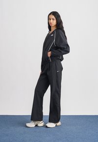 Fekete sportos melegítő, cipzáras felsővel, fehér csíkozással és nadrággal. Bézs sportcipővel viselve, semleges háttér előtt bemutatva.