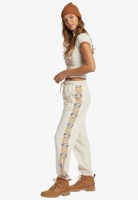 Pantalones jogger color crema con una franja lateral estampada en marrón y dorado, combinados con un top corto color beige y botas marrones con cordones. Gorro de punto texturizado.