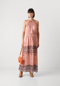 Robe maxi rose avec des motifs floraux et cachemire, décolleté croisé, taille froncée, et ourlet à motifs contrastés. Bottes ankle marron et sac orange.
