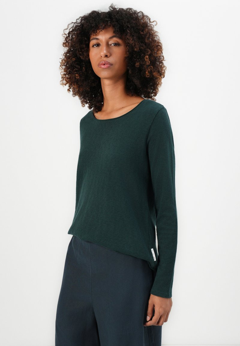 Dunkelgrünes Langarmshirt mit Rundhalsausschnitt, weichem Material und asymmetrischem Saum. Kombiniert mit dunklen Hosen für einen Kontrast.