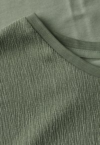 Texturerad grön tyg med en ribbad halslinning och kontrasterande slät sektion. Materialet verkar lätt med en subtil lyster.