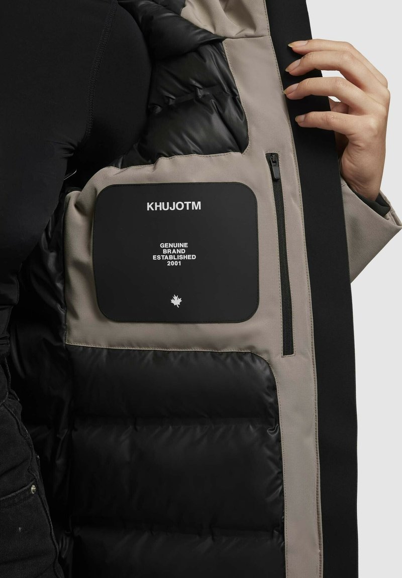 Interno di un giubbotto imbottito nero con fodera beige. Presenta un grande etichetta nera con testo bianco e una tasca con zip. Superfici lisce e testurizzate.